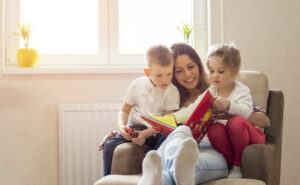 Mother reading book in comfy chair with son and daughter with sunny window in background